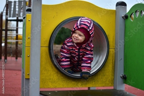 Fototapeta Smiling toddler in a colorful striped snowsuit playing inside a playground tunnel on an autumn day. Bright yellow structure and soft light capture a joyful, candid childhood moment.