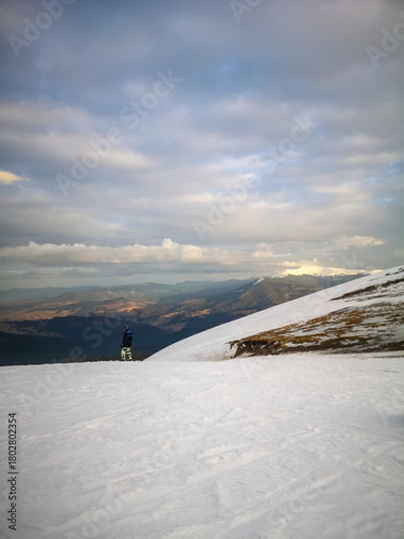 Fototapeta Person with snowboard standing on snowy mountain slope looking at wide winter landscape with soft clouds and warm sunset tones in vertical format.