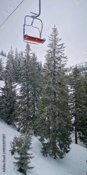 Fototapeta Empty ski lift chair gliding above frosted pine trees on a snowy mountain slope under a pale winter sky, capturing a quiet alpine atmosphere.