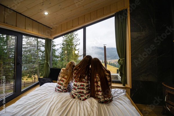 Obraz Family in matching pajamas admiring Mountain View. Three girls in festive Christmas pajamas sitting on a bed, peacefully looking out large cabin windows at the scenic mountain landscape.