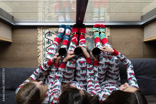 Obraz Top view of family in pajamas with mugs. Three girls wearing festive pajamas and colorful socks hold mugs while relaxing indoors, seen from above near a glass window.