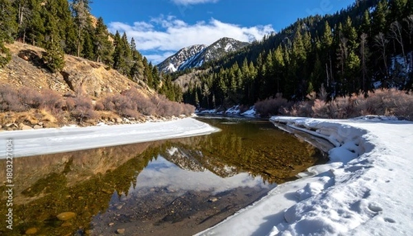 Fototapeta A serene river flows between snow-covered banks and forested hills under a blue sky, reflecting the surrounding landscape