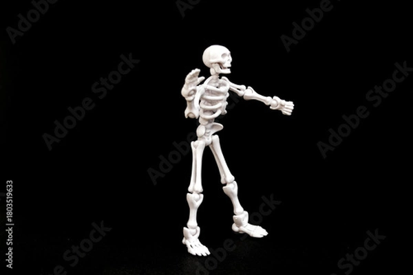 Fototapeta A small, articulated human skeleton model with red eyes, standing upright on a stark black background. Concepts of anatomy, biology, Halloween, fear, science, education, medical, spooky, and death.