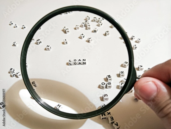 Fototapeta A magnifying glass highlights the word “LOAN” among scattered letter cubes, symbolizing loan review, financial assessment, and careful evaluation in banking or credit decisions.