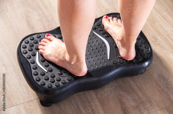Fototapeta Women's legs stand on a sports vibration platform at home. Concept of doing sports at home. Modern home exercise machine, close-up