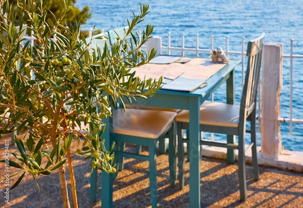 Fototapeta Sithonia, Chalkidiki, Greece. Traditional tavern cafe exterior with wooden table chairs. Olive tree branch focused in front with the view of the aegean sea in the background..