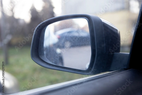 Fototapeta Car side mirror reflecting parking lot with cars and greenery in the background during daytime