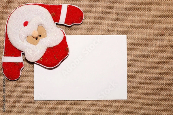 Fototapeta A letter to the santa. Toy Santa holds a white envelope in his hands with a letter in the background of a sackcloth