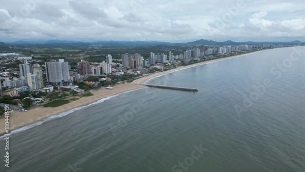 Obraz Aerial shot of a long pier jutting into a calm, gray sea under a cloudy sky. Unidentifiable people stand at the end, with distant islands on the horizon. Peaceful, minimalist coastal scene.