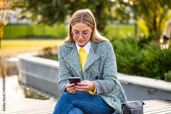 Fototapeta Portrait of urban businesswoman with eyeglasses in stylish coat using smartphone while sitting on park bench in the city during autumn day.	