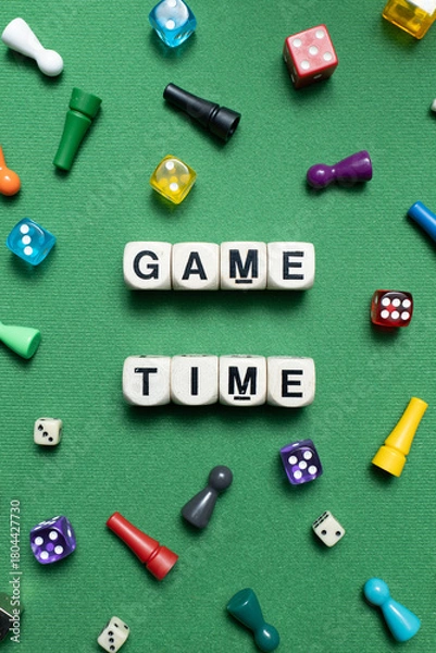 Obraz Wooden dice show the phrase 'Game Time' with a selection of colourful dice and game tokens lying around it on a vibrant green baize fabric