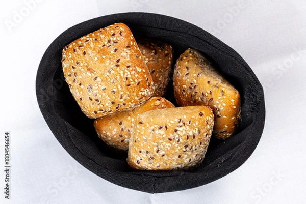 Fototapeta Four fresh whole-grain bread rolls covered with sesame and flax seeds, served in a black bread basket on a white tablecloth.