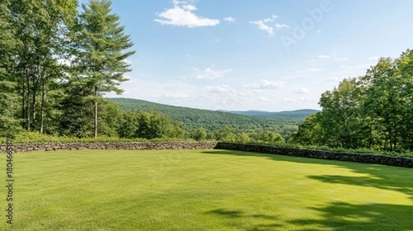 Fototapeta Expansive green lawn bordered by a stone wall leads to rolling hills and a bright blue sky