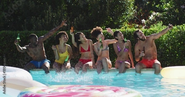 Fototapeta Clinking bottles, six friends sitting on backyard pool edge in swimwear, splashing near donut float