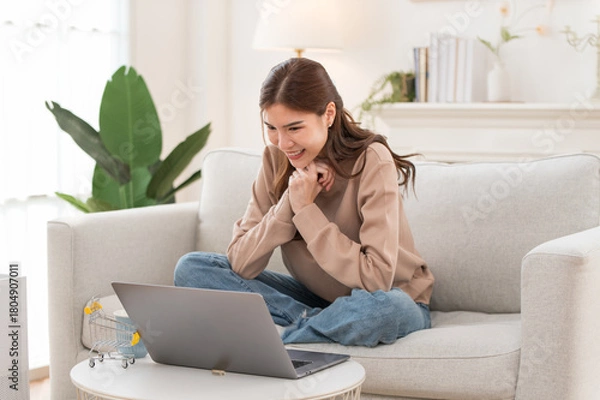 Obraz Concentrated Asian girl sit on couch working on laptop browsing internet at home, young woman busy using computer surfing wireless web shopping online
