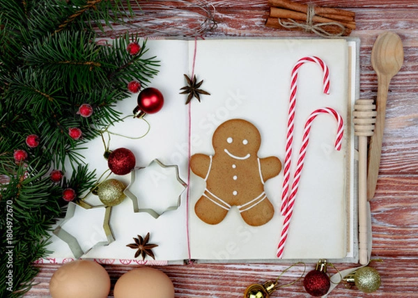 Fototapeta Christmas festive flatlay composition with vintage old recipe book, gingerbread man and Christmas decorations on the wooden background. Homemade holiday baking. Close-up. Selective focus.