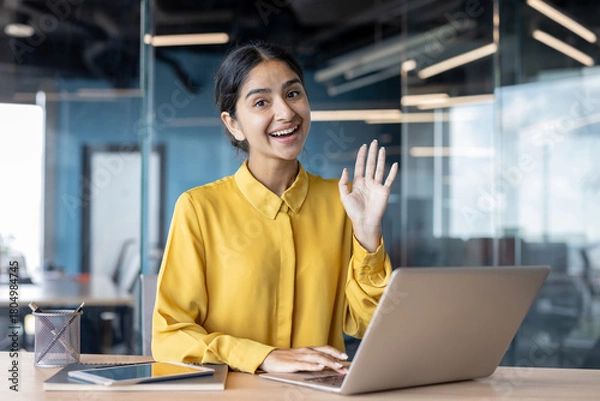 Fototapeta Portrait of a smiling young Indian woman sitting at an office desk, working on a laptop, greeting and waving at the camera