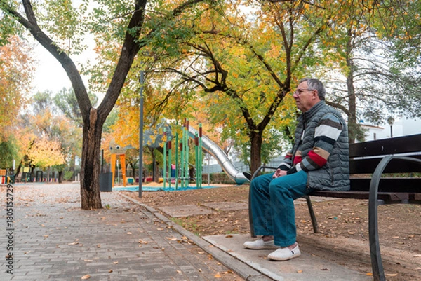 Fototapeta An older man sits on a park bench near a playground, enjoying a quiet break amid colorful autumn trees and soft natural light.