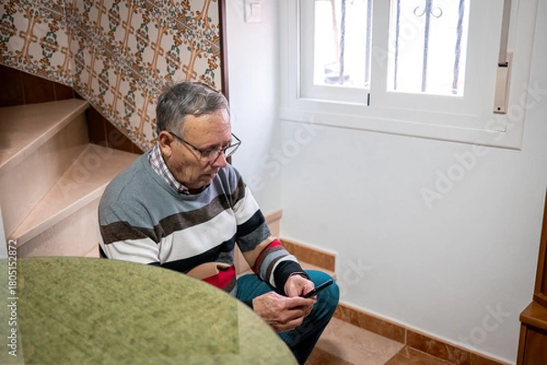 Fototapeta Elderly man sits by the stairs inside his home, focused on his smartphone, in a calm domestic environment with natural light from a nearby window.