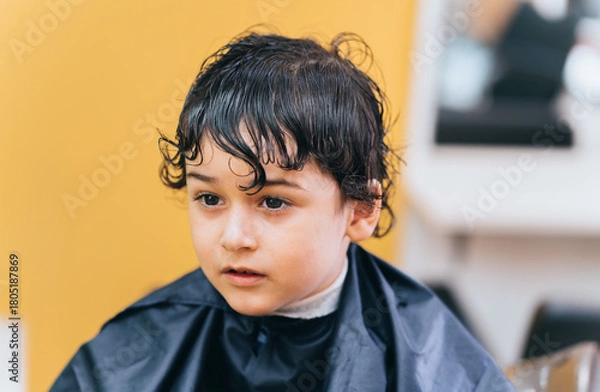 Fototapeta Young child with wet hair sits in a salon with a navy cape, looking thoughtfully ahead after a haircut.