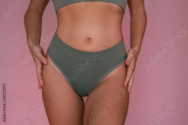 Fototapeta Young woman touching her hips in minimalistic green underwear, showing smooth skin after laser hair removal and feminine curves, standing against a light pink background