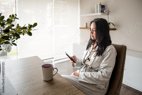 Fototapeta A woman is sitting at a table with a cell phone in her hand