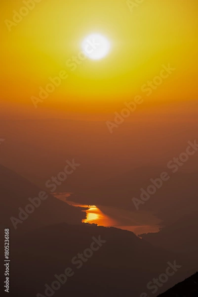 Obraz Brilliant morning sun hangs high in a golden sky, its intense light casting shimmering, molten gold reflection upon winding river nestled deep within hazy, silhouetted valley as viewed from Dobratsch