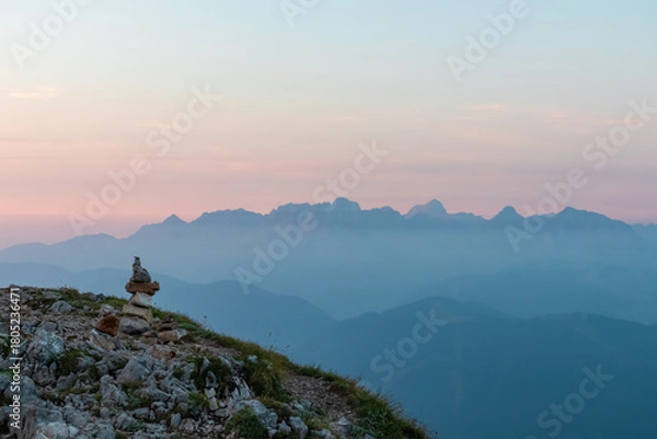 Fototapeta Stone cairn marks a hiking trail on a rugged mountaintop, guiding the way towards a magnificent panoramic view of distant, mist-covered mountain ranges under a serene and soft-colored pastel dawn sky.