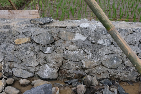 Fototapeta A close-up view of a house foundation made from stacked natural stones and cement, showing the rough texture and early construction stage on a muddy worksite.