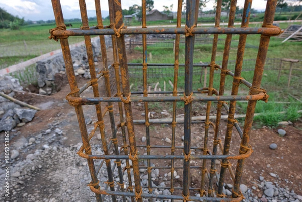 Obraz A close-up view of a reinforced steel column frame partially covered in fresh concrete at a house construction site, showing the early stage of foundation and structural work.