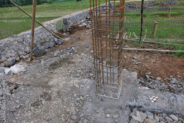 Fototapeta A close-up view of a reinforced steel column frame partially covered in fresh concrete at a house construction site, showing the early stage of foundation and structural work.