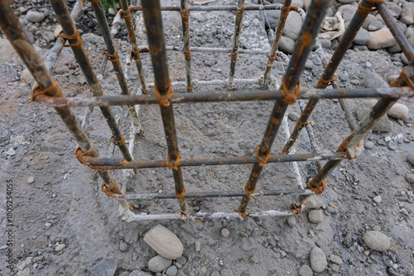Fototapeta A close-up view of a reinforced steel column frame partially covered in fresh concrete at a house construction site, showing the early stage of foundation and structural work.