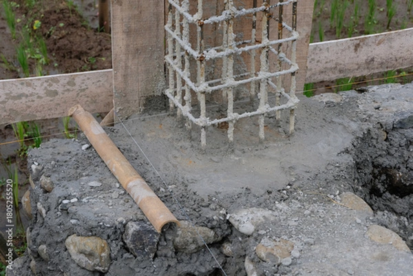 Fototapeta A close-up view of a reinforced steel column frame partially covered in fresh concrete at a house construction site, showing the early stage of foundation and structural work.