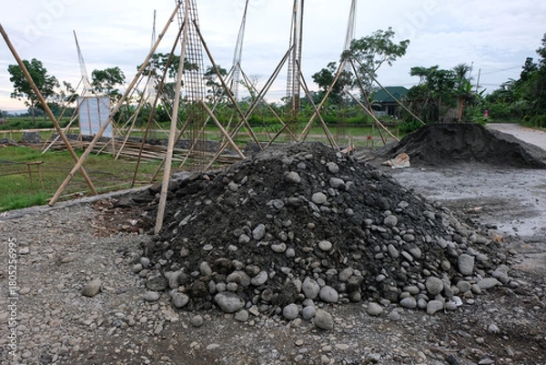 Fototapeta A pile of construction materials, including gravel and soil, at a house-building site with reinforced steel columns and wooden supports visible in the background.

