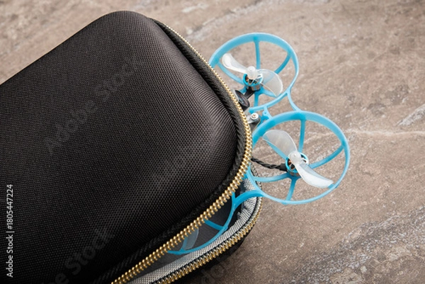Fototapeta compact black zipper case partially holds a small FPV "Tiny Whoop" drone, with its two bright blue ducted propellers. quadcopter represents the beginner equipment for learning FPV flying.