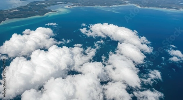 Obraz Aerial view of a turquoise ocean with puffy white clouds below, island coastline visible on the horizon, serene & vast