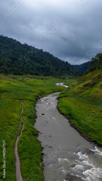 Fototapeta Clear mountain water flows over rocks in a vibrant green valley, surrounded by majestic peaks at dawn.