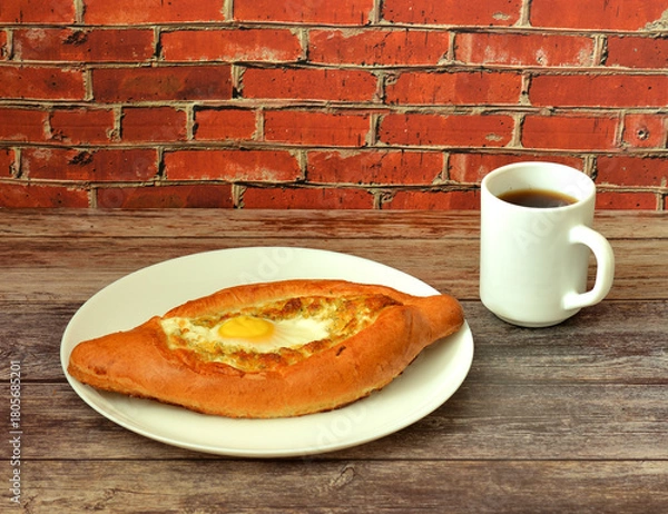 Fototapeta Khachapuri, traditional Georgian pie with egg and cheese in a plate on a wooden table, next to a cup of hot tea.