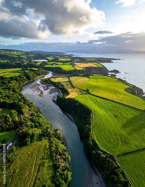 Fototapeta Aerial view of a river winding through vibrant green fields toward a sunlit coast under a cloudy sky