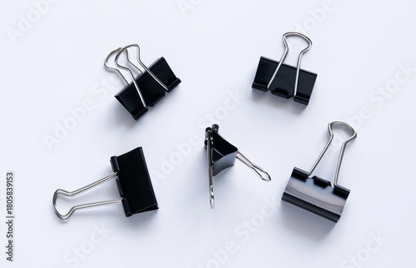 Fototapeta Five black binder clips scattered on a white background, highlighting their metal handles and dark color.