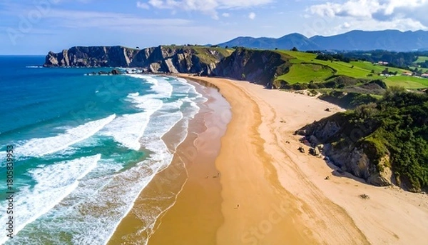 Fototapeta Aerial view of a wide, sandy beach with blue waves, rocky cliffs, and green hills under a partly cloudy sky