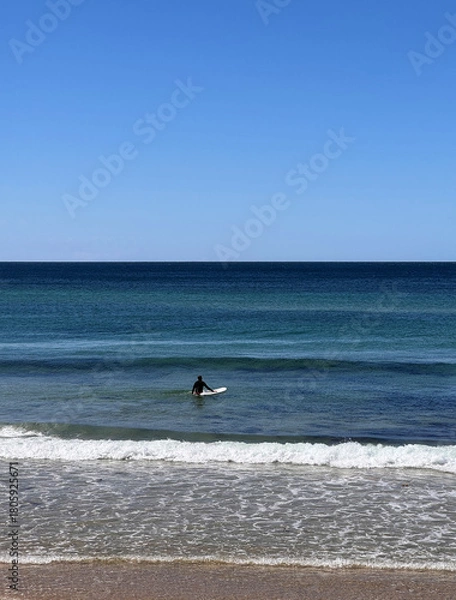 Obraz rear view of man wearing wetsuit entering ocean with surfboard wading through water going for a surf on a sunny day against clear blue sky with space for text