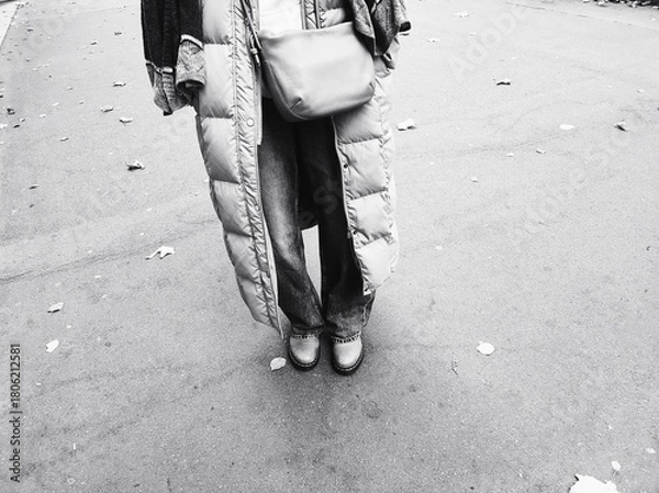 Fototapeta Street Style in Black and White. A person wearing a long puffer coat stands alone on a street, capturing a moment of everyday urban fashion. The monochrome palette adds a timeless feel.