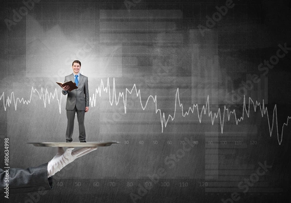 Fototapeta Businessman on metal tray with red book in hands against graphs background