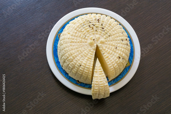 Obraz Вelicious round white homemade cheese with textured, ridged surface and slice cut out, placed on decorative plate with blue and gold rim, revealing its creamy interior