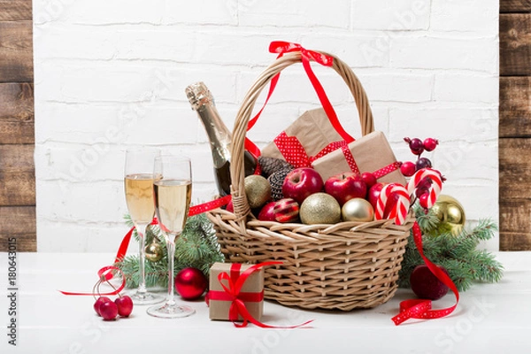 Fototapeta Two glasses of champagne with Christmas basket with bottle, gifts with red satin ribbon, candy canes, pine cones, golden garlands on white brick wall background, new year celebration concept