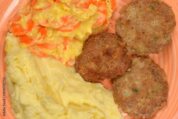 Fototapeta A close-up, top-down view shows three browned meat patties, creamy mashed potatoes, and carrots in a light sauce on a peach-colored plate