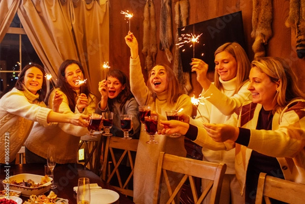 Fototapeta Young women in knitted sweaters laugh and raise glasses of mulled wine. Friends have fun and drink wine with spices. New Year Eve party with sparklers.