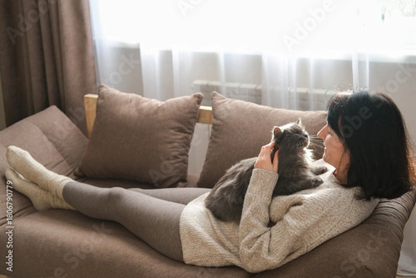 Fototapeta Smiling woman is lying on brown sofa, stroking at fluffy gray cat resting on her chest, bathed in window sunlight. Pet companionship, relaxation, cozy home life, love for animals