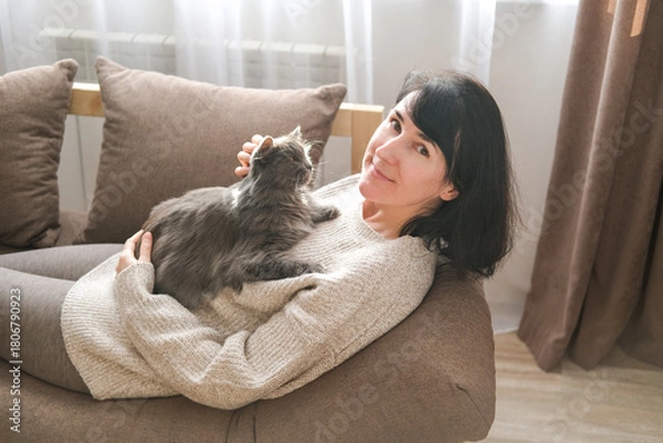 Obraz Smiling woman is wearing cozy sweater is lying on sofa, holding fluffy gray cat on her chest and looking calmly at camera in brightly lit room. Pet companionship, relaxation, affection with animals